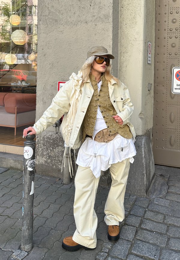 Persoon met een beige pet, oversized zonnebril, gelaagde beige en witte kleding en bruine schoenen leunend tegen een paaltje op een stadsstoep.