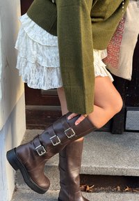 Personne portant un pull vert, une jupe en dentelle blanche et de grandes bottes marron à boucles, ajustant une botte en se tenant sur une marche à l'extérieur.