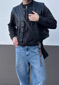 Μαύρο δερμάτινο γιλέκο πάνω από ένα μαύρο πουλόβερ, συνδυασμένο με ανοιχτό μπλε τζιν. Περιλαμβάνει κουμπιά, φερμουάρ και αξεσουάρ γαντζιού.