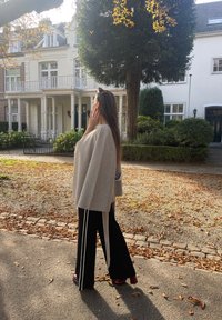 Γκρι μάλλινο κασκόλ με χαλαρή εφαρμογή, συνδυασμένο με μαύρα παντελόνια με φαρδιά πόδια που έχουν λευκές λωρίδες στο πλάι. Μακρι ά καστανά μαλλιά κυλούν πίσω.