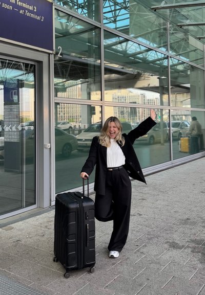 Mujer sonriente con traje negro sosteniendo una maleta negra con ruedas, levantando un brazo, de pie frente a una gran terminal de aeropuerto de cristal con coches al fondo.