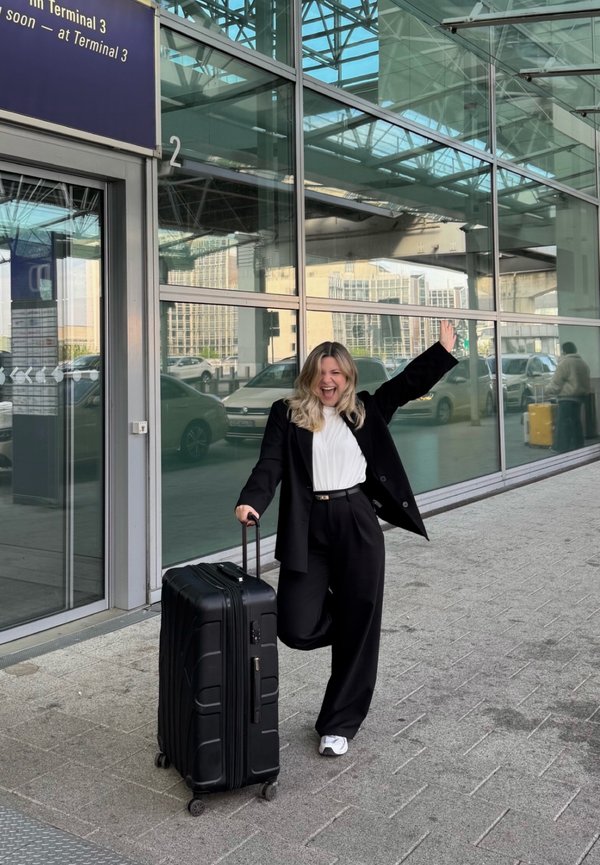Usmievavá žena v čiernom obleku drží čierny kufor na kolieskach, zdvíha jednu ruku, stojí pred veľkým skleneným letiskovým terminálom s autami v pozadí.