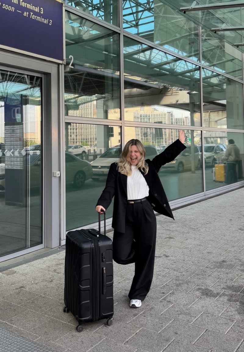 Femme souriante en costume noir tenant une valise noire à roulettes, levant un bras, debout devant un grand terminal d'aéroport en verre avec des voitures en arrière-plan.