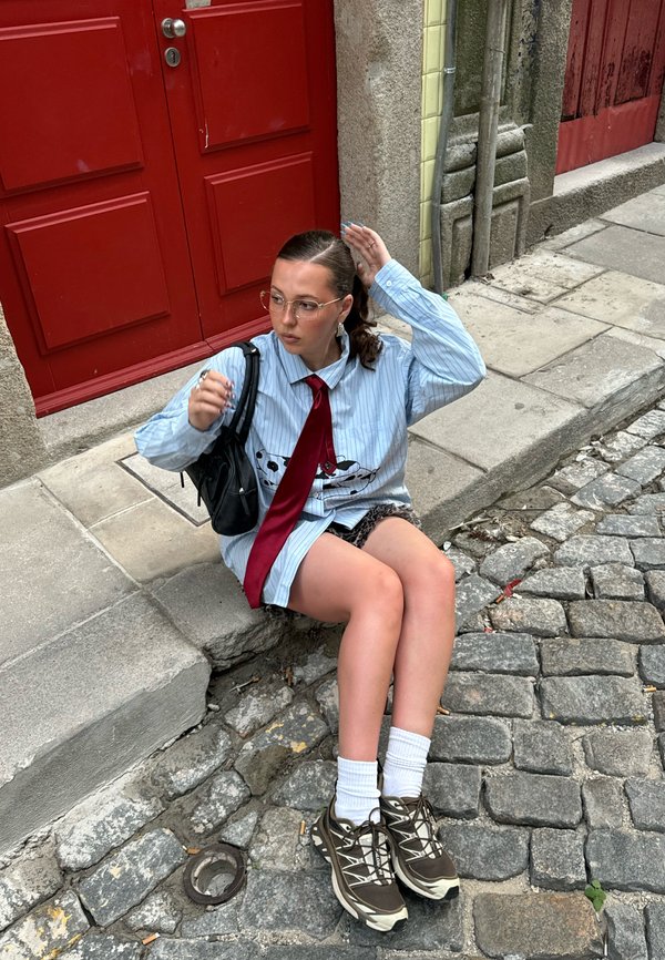 Túlméretezett kék csíkos ing grafikai mintával, piros nyakkendő, fekete kézitáska, mintás rövidnadrág, fehér zokni és barna tornacipő macskaköves utcán.
