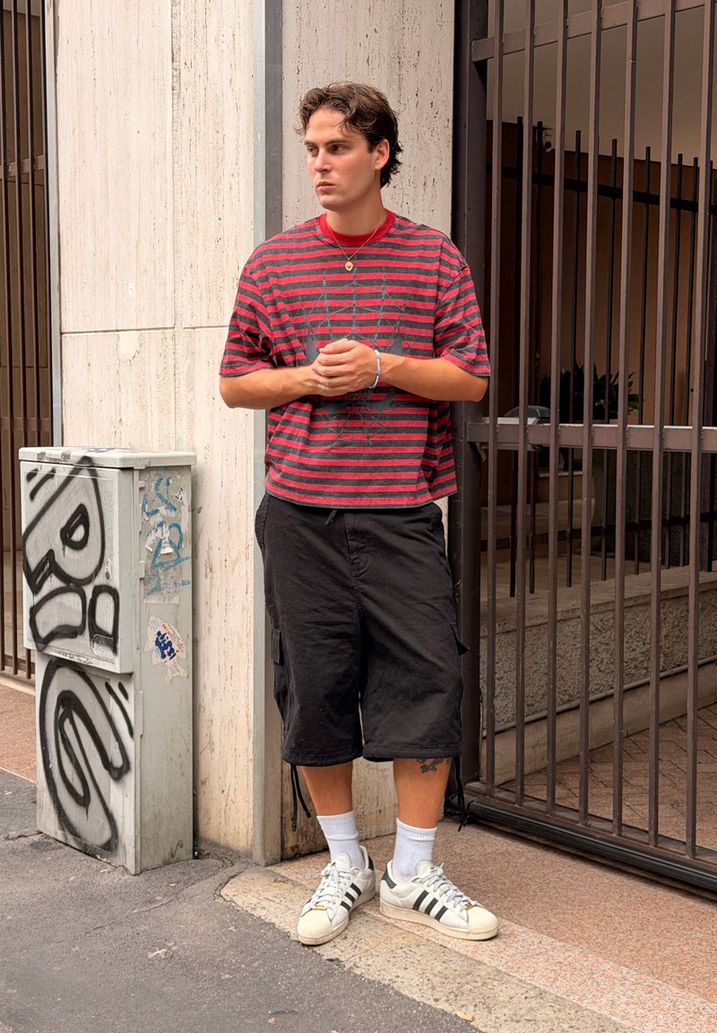 Jeune homme portant une chemise rayée rouge et noire et un short noir, debout près d'une entrée grillagée sur un trottoir urbain avec des graffitis sur une armoire technique.