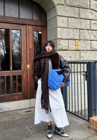 Pehmendusega sinine kott, pruun sall, nahktagi, hall top, valge seelik, mustad sokid ja mustriga kingad. Seisab puust uste lähedal.