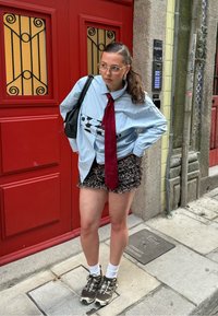 Túlméretezett kék csíkos ing fekete-fehér grafikával, bordó nyakkendő, fodros leopárdmintás rövidnadrág, fehér zokni és sötét sportcipő. Piros ajtó háttér.