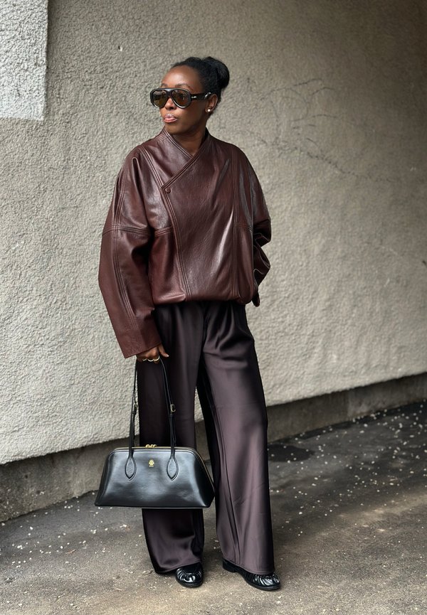 Kvinde iført oversized brun læderjakke, vide mørke bukser, sorte loafers, store solbriller, holder en sort håndtaske, står ved en tekstureret væg.