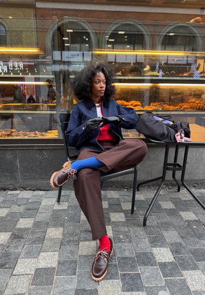 Joven sentada con las piernas cruzadas, lleva calcetines y guantes coloridos, en la mesa de una cafetería al aire libre frente a una vitrina de panadería.