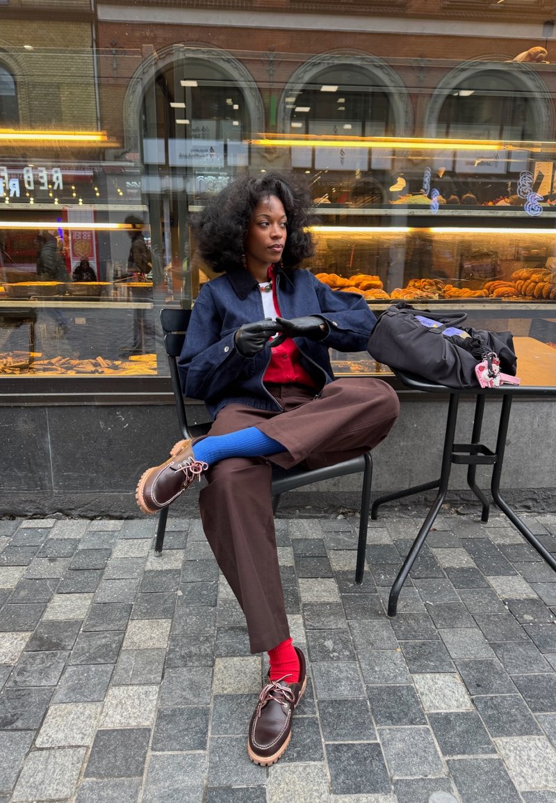 Giovane donna con calzini e guanti colorati seduta a gambe incrociate al tavolo di un caffè all'aperto davanti alla vetrina di una panetteria.