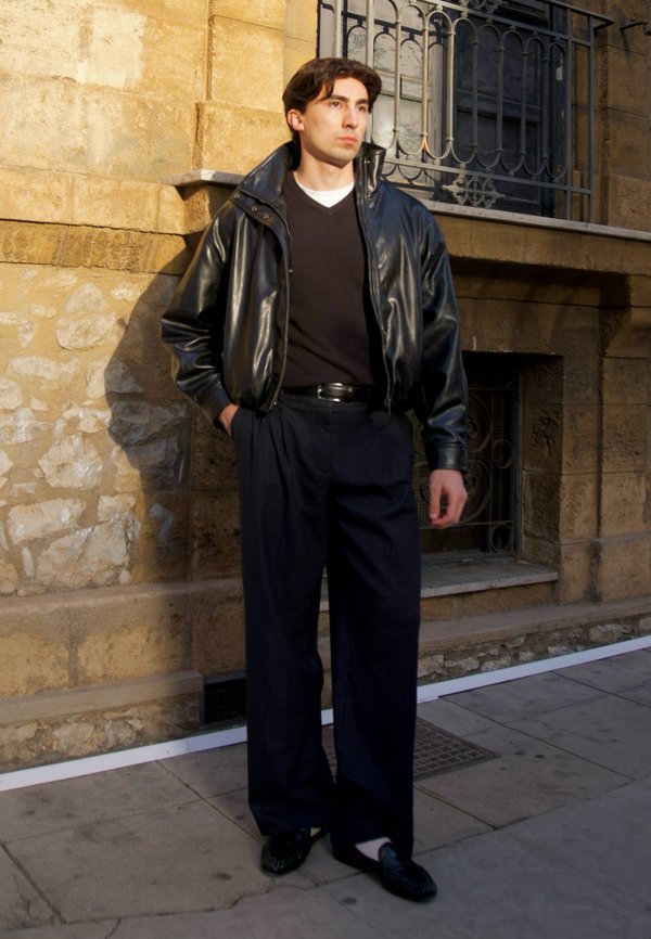 Junger Mann trägt eine schwarze Lederjacke, schwarzen Pullover, dunkle, weite Hose und Loafers und steht an einer Steinmauer eines Gebäudes.