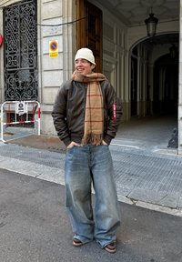 Ruda odinė bomber tipo striukė, smėlio spalvos šalikas su raudonomis juostelėmis, plačios mėlynos džinsai ir rudi batai. Aplinka apima architektūrinius elementus ir ženklinimą.