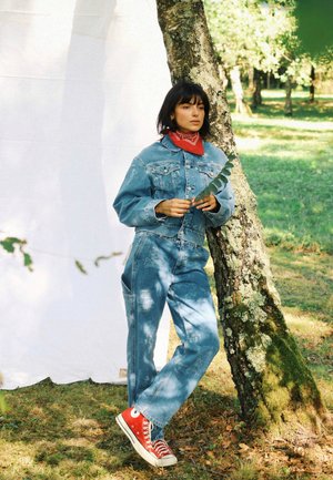 Jeansjacke und Latzhose in Hellblau mit verblassten Mustern. Rotes Halstuch um den Hals und rote High-Top-Sneaker. Grüne Blätter in der Hand gehalten.