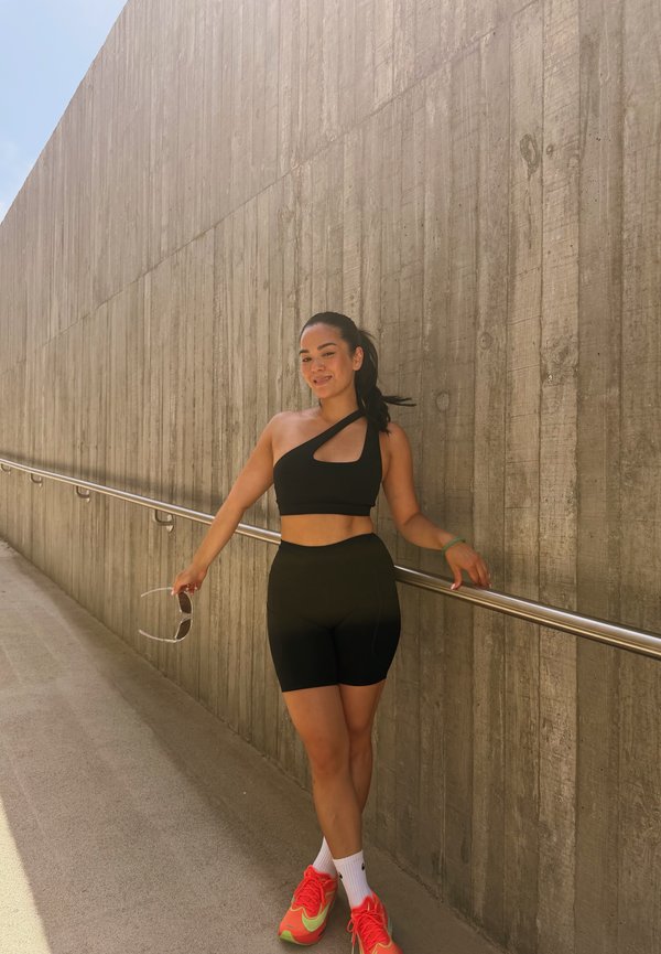 Jeune femme en tenue de sport noire et baskets éclatantes, debout près d’un mur en béton, tenant des lunettes de soleil et souriant au soleil extérieur.