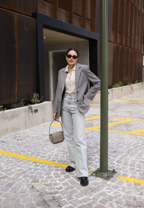 Tjekket blazer, beige bluse, lyseblå bukser med vide ben, sorte sko, grå håndtaske; stående mod en struktureret stengulv baggrund.