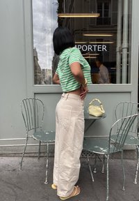 Grün-weißer gestreifter Cropped-Shirt, hellbeige weit geschnittene Hose mit aufgenähten Taschen, gelbe Handtasche und rosa Flip-Flops. Außenbereich.