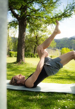 Una persona se encuentra tumbada sobre una esterilla de yoga gris claro en la hierba, realizando un ejercicio, vestida con una camiseta sin mangas negra y pantalones cortos grises.