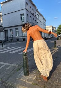 Beige, vloeiende broeken met een elastische tailleband en een gestructureerde afwerking. De figuur staat naast een groene metalen paal op een straat van kinderkopjes.