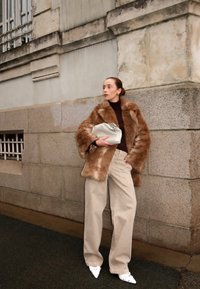 Bruine nepbontjas, beige wijde broeken, witte puntige schoenen, met een crèmekleurige clutch. Gestructureerde stof met zachte accenten.