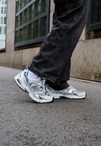Fehér és szürke New Balance tornacipők, hálós és gumírozott anyagból, kiemelkedő "N" logóval és fekete részletekkel. Sötét nadrággal viselve.