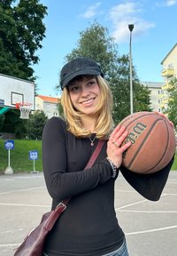 En basketboll med en brun, texturerad yta hålls av en person i en svart långärmad tröja, svart keps och en vinröd väska i en spelplats.