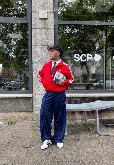 Chaqueta de pista roja de Adidas con rayas blancas, pantalones de pista navy y zapatillas blancas. Sosteniendo un libro con una cubierta azul, de pie cerca de un escaparate de cristal.