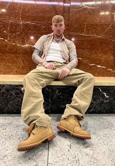 Botas de senderismo marrones con suelas de goma, pantalones cargo beiges y una camisa de cuadros corta sobre una camiseta blanca, sentado en un banco de piedra frente a un fondo de mármol.