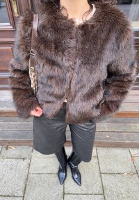 Personne portant une veste en fourrure marron, une jupe en cuir noire longueur genou, des bottes noires pointues et portant un sac à bandoulière marron, debout en plein air.