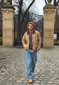 Veste bomber beige avec col marron, pull gris et jean bleu ample. Chaussures marron clair. Pavé en pierre et porte métallique en arrière-plan.