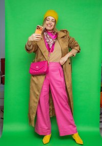 Femme souriante portant un bonnet jaune, un manteau marron, un pantalon rose et des bottes jaunes, prenant un selfie devant un fond vert, tenant un sac à main rose.