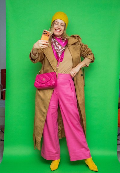 Mujer sonriente con gorro amarillo, abrigo marrón, pantalones rosas y botas amarillas, se toma una selfie frente a un fondo verde, sosteniendo un bolso rosa.