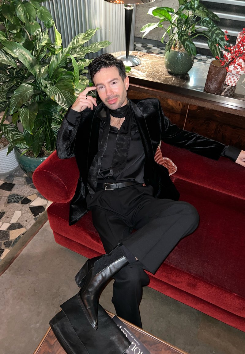 Blazer in velluto nero, camicia nera con sciarpa, pantaloni neri aderenti e stivali neri a punta, seduto su un divano rosso con piante sullo sfondo.