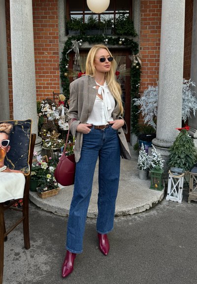 Blazer de cuadros marrón sobre una blusa blanca con un detalle de lazo, jeans de tiro alto azules y botas de tobillo burdeos, combinados con un bolso rojo.