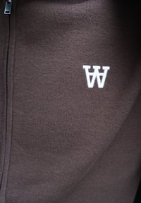 Καφέ πλεκτό ύφασμα με κάθετο φερμουάρ και λευκό κεντημένο λογότυπο που μοιάζει με δύο αναποδογυρισμένα γράμματα W στη δεξιά πλευρά.