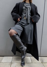 Femme portant un manteau noir, un pull gris avec un nœud, une jupe grise à fines rayures et des bottes noires montantes, debout contre un mur gris.