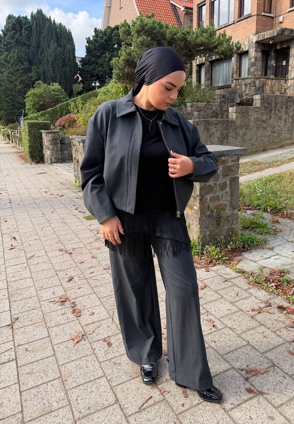 Une femme portant un foulard noir, une veste grise et un pantalon à jambes larges se tient sur un trottoir, près d’un mur en pierre et de maisons en briques.