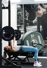 En skivstång med vikter som lyfts av en person på en svart viktbänk. Personen bär en vit crop top och blå mönstrade leggings. Inomhusgymmiljö.