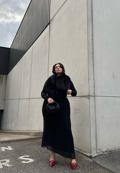 Vestido largo de encaje negro con una silueta ajustada, stole de piel texturizada, bolso negro y tacones rojos de punta en un fondo de concreto.