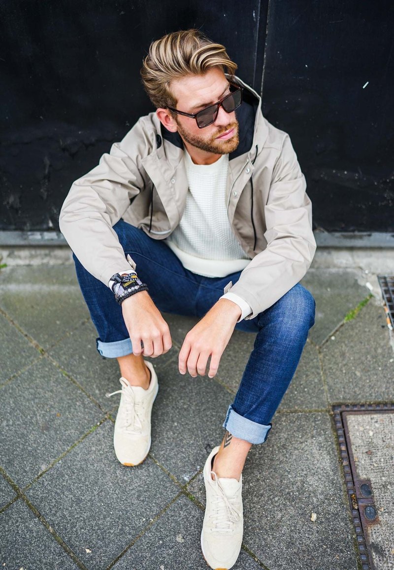 Beige jacket with black accents over a white sweater, dark jeans, and light sneakers, featuring rolled cuffs and a bracelet on the wrist.
