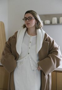 Jauna sieviete ar brillēm, valkājot baltu pogājamu kardiganu un brūnu p�ārmēru mēteli, stāv iekštelpās ar rokām uz gurniem.