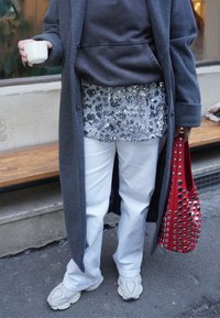 Manteau gris, sweat à capuche noir, haut en sequins argentés, pantalon blanc et baskets beige. Sac rouge avec découpes circulaires. Sol et banc en bois visibles.