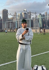 Sweat-shirt gris oversize et pantalon de survêtement assorti, avec une ceinture noire. Un bonnet vert arbore un logo. Un homme tient un smartphone sur un terrain herbeux.