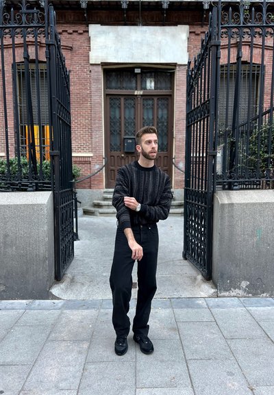 Suéter negro con estampado y mangas largas, combinado con pantalones negros y zapatillas deportivas. Escenario al aire libre con paredes de ladrillo y puertas de hierro forjado.