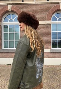 Olijfgroene leren jack met een draak grafische print op de achterkant, gestructureerde stof, en een bruine bontmuts. Laat een relaxte pasvorm en details zien.