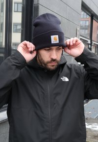 Un homme ajuste un bonnet noir Carhartt tout en portant une veste noire The North Face à l'extérieur d'un bâtiment urbain par une journée nuageuse.