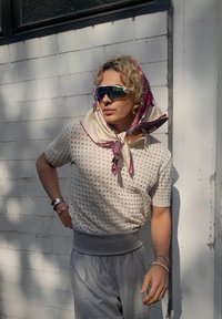 Kötött, törtfehér, rövid ujjú felső, texturált lyukakkal, szürke derékkal, mintás selyemsál és tükröződő napszemüveg kíséretében. Szürke nadrág.