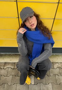 Jovem usando um boné cinza e um suéter, cachecol azul, handbag preto e ténis amarelos, sentado contra um fundo amarelo.