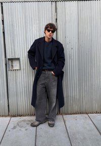 Long manteau bleu marine, pull foncé, et jean gris à jambes larges, assortis de chaussures grises. Entouré d'un fond en métal ondulé.