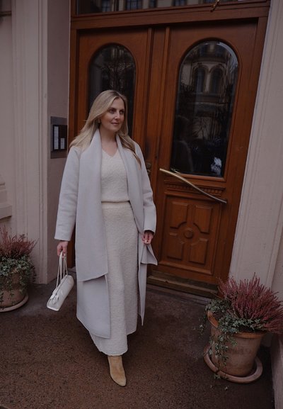 Abrigo de lana gris claro sobre un vestido texturizado crema, combinado con botas de tobillo beige y un pequeño bolso blanco, todo frente a puertas de madera.