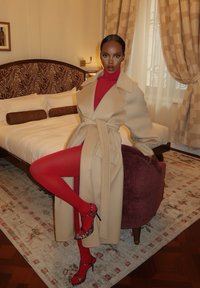 Abrigo de lana beige con cinturón anudado, jersey de cuello alto rojo y medias rojas. Combinado con zapatos de tacón de estampado de leopardo, posando en una habitación de hotel con cama y alfombra estampada.